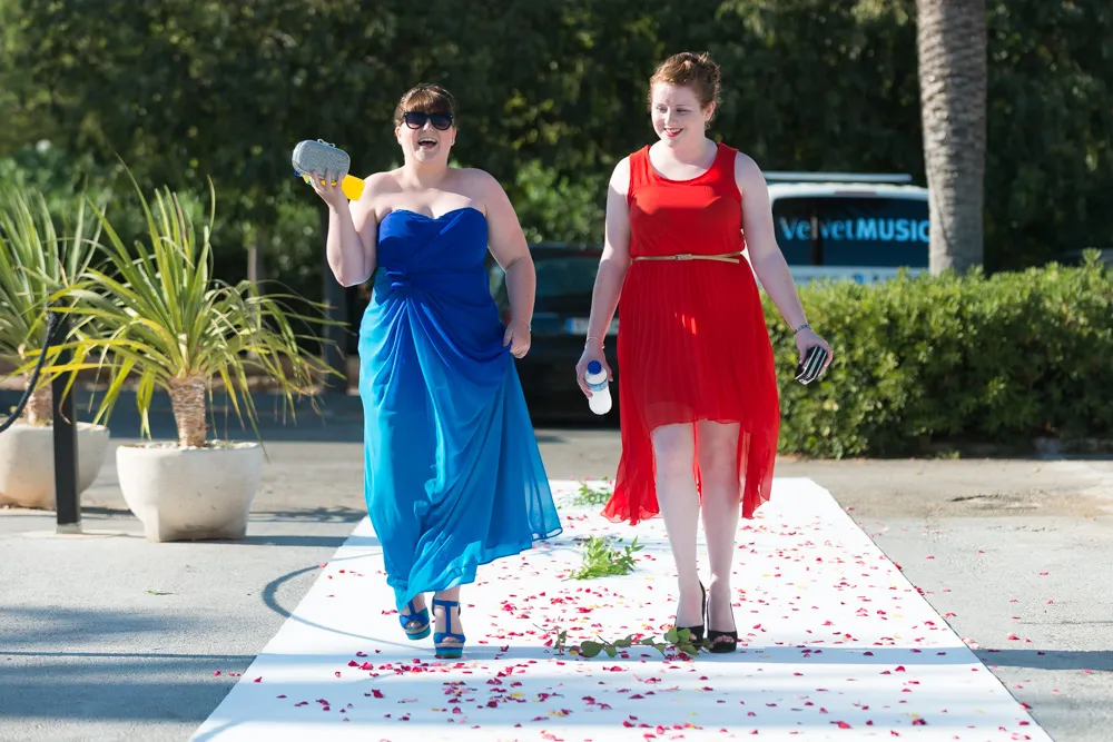 Two women walking on a white aisle covered with rose petals; one in a blue dress holding a clutch, the other in a red dress holding a water bottle and phone.