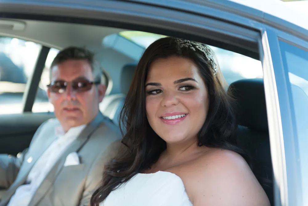 Smiling bride with dark hair and a tiara sitting in a car next to a man wearing sunglasses and a light suit.
