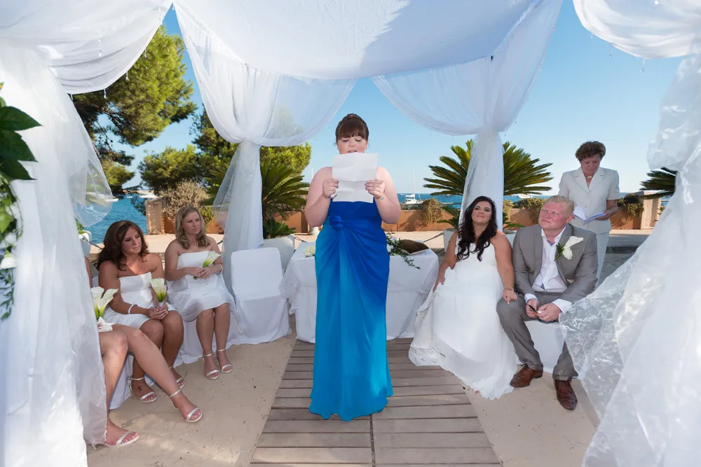 Woman in blue dress reading a speech at a beach wedding ceremony under white draped canopy with bride, groom, and guests seated.