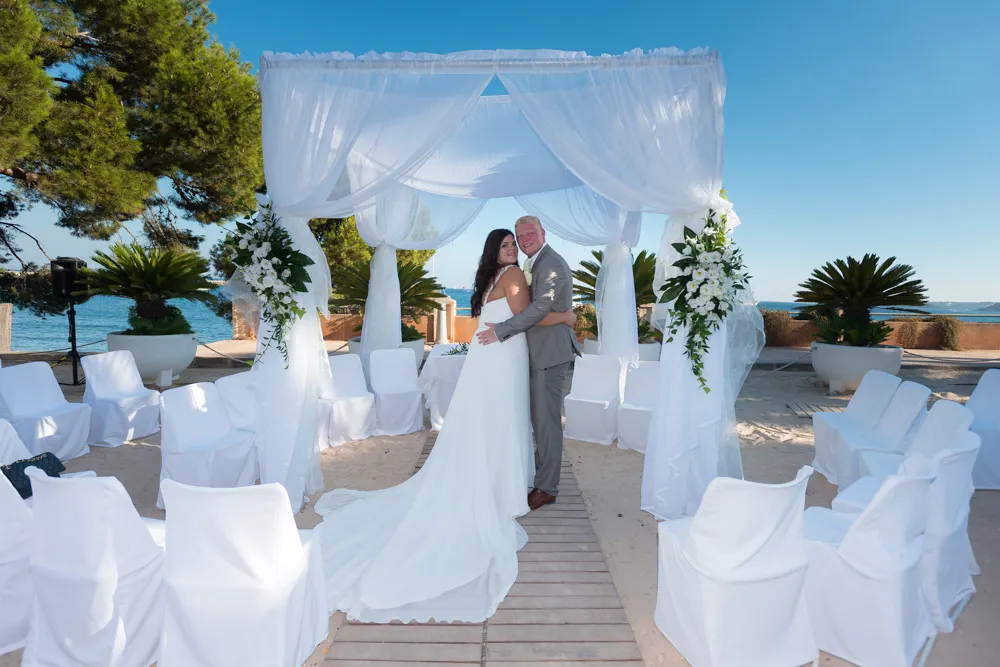 Bride and groom embracing under white draped wedding arch on a beach with white covered chairs arranged around.