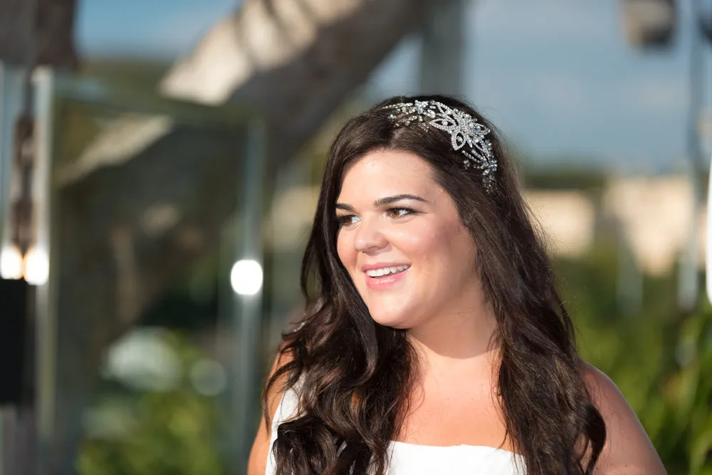 Smiling woman with long dark hair wearing a white dress and a jeweled hair accessory outdoors.