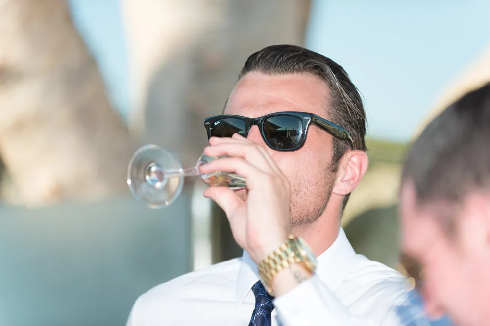 Man in sunglasses and white shirt drinking from a wine glass outdoors during the day.