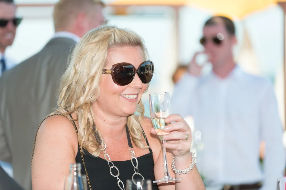 Smiling woman with blonde hair wearing large sunglasses and a black dress, holding a glass of champagne at a social event.