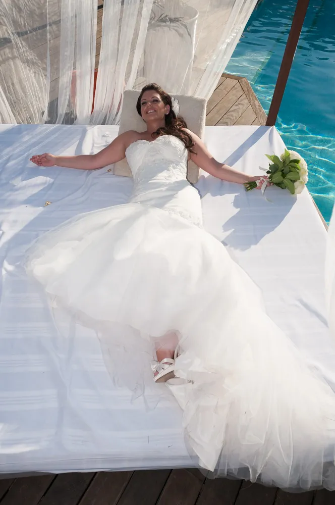 Bride in white wedding dress lying on a sunbed by a pool, smiling and holding a bouquet.