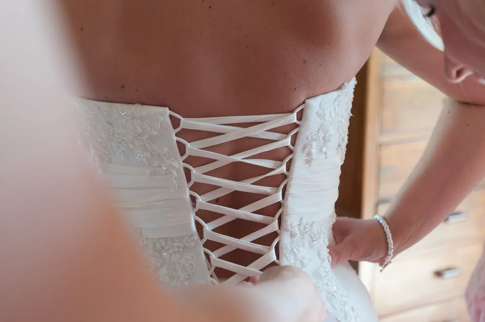 Close-up of a bride's back as someone laces up the corset-style back of her white wedding dress with lace and bead details.