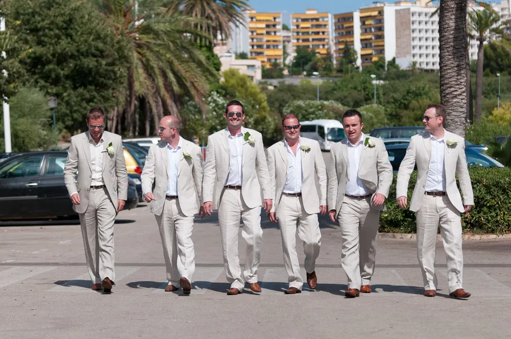 Six men in matching light beige suits, white shirts, and brown shoes walking outdoors on a sunny day with palm trees and buildings in the background.