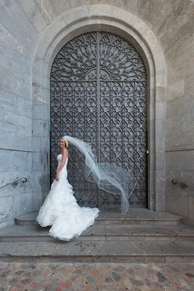 Bride in a strapless white wedding gown with a long flowing veil standing on stone steps in front of a large ornate iron door.