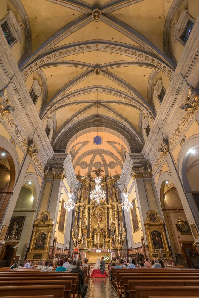 Interior view of a church during a wedding ceremony with guests seated and a couple kneeling at the altar under ornate arches and chandeliers.