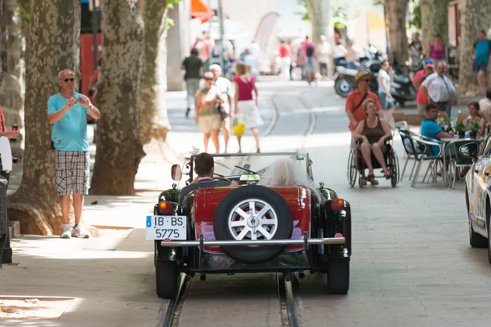 Rear view of a vintage convertible car driving on a street with tram tracks, carrying a bride and groom, surrounded by pedestrians and people sitting at outdoor tables.