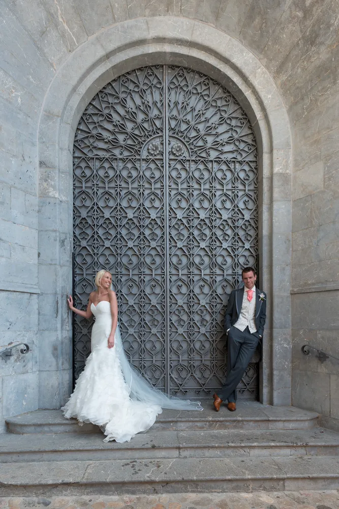 Bride in white gown and groom in suit posing separately against a large ornate arched metal door.