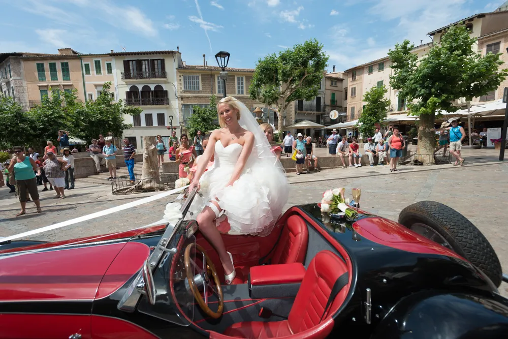 Smiling bride in a white dress sitting on the door of a red and black vintage convertible car in a sunlit town square with onlookers in the background.