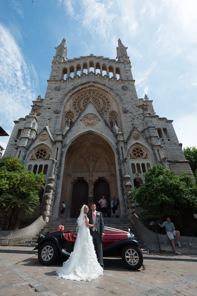 Bride and groom posing in front of a vintage car outside a large, ornate stone church with an arched entrance.