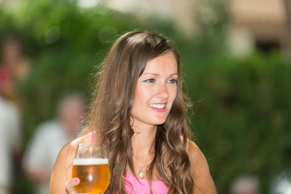 Smiling woman with long brown hair wearing a pink top and holding a glass of beer outdoors.