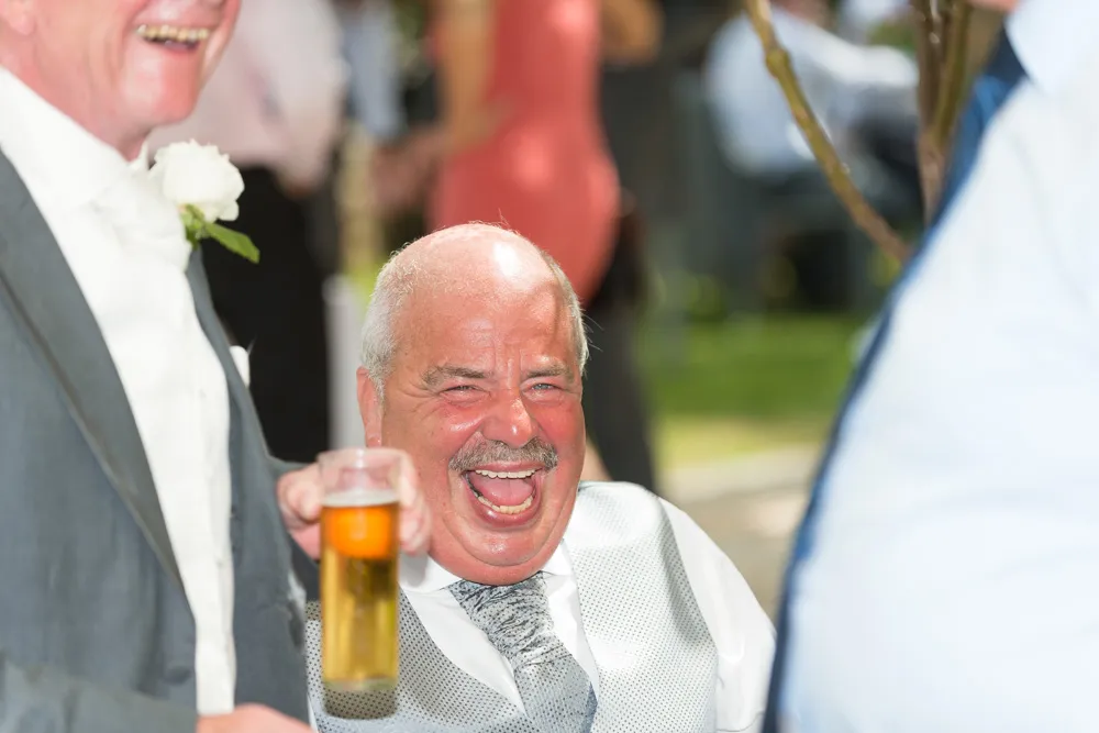 Smiling older man in a silver waistcoat laughing joyfully while holding a glass of beer at an outdoor event.