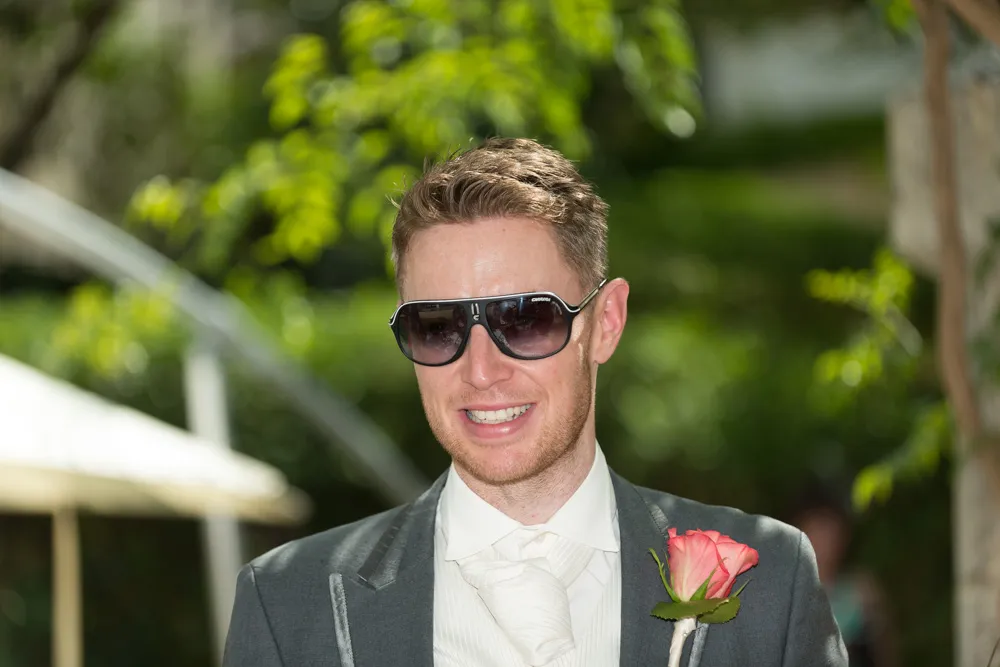 Smiling man wearing sunglasses, a gray suit, white shirt, and tie with a pink rose boutonniere outdoors.