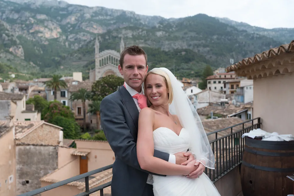 Smiling bride and groom embracing on a balcony with a mountainous townscape in the background.