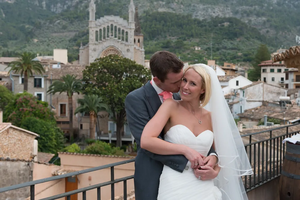 Bride in a white dress and veil smiling while groom embraces and kisses her on a balcony with a historic town and church in the background.