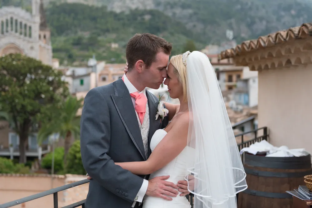 Bride and groom embracing and touching foreheads on a balcony overlooking a village with mountains in the background.