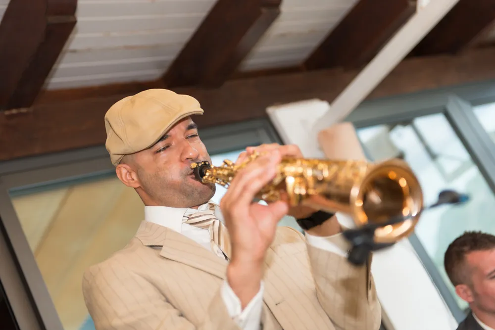 Man wearing beige suit and cap playing a saxophone indoors.