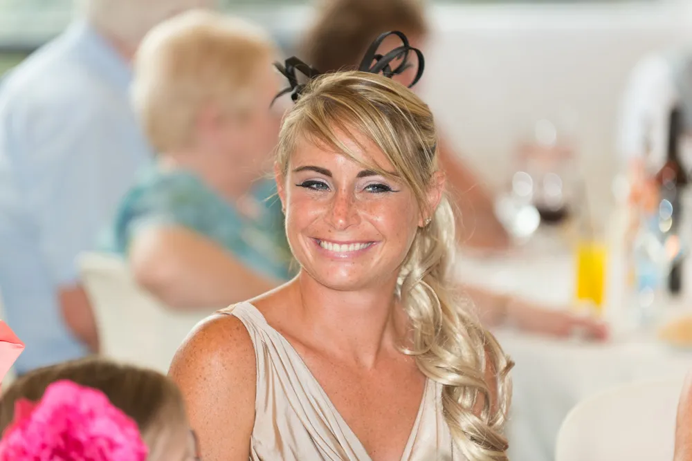 Smiling woman with long blonde hair styled to the side and a black fascinator, wearing a beige dress at an indoor event.