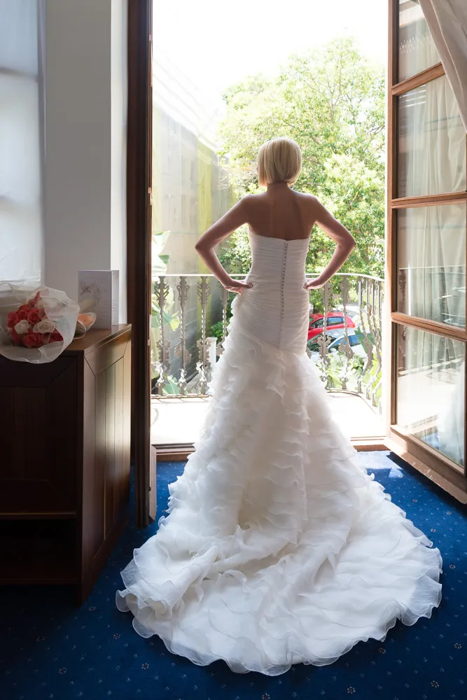 Bride in a strapless white wedding gown standing with hands on hips, facing an open balcony with greenery outside.