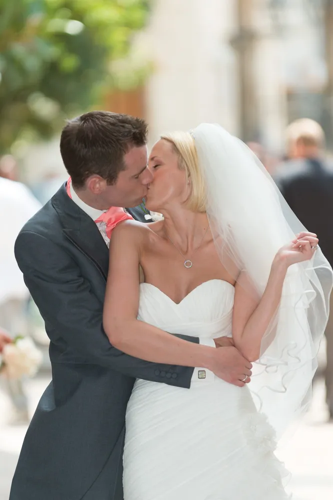 Bride and groom sharing a kiss outdoors, the bride wearing a white dress and veil, and the groom in a dark suit.