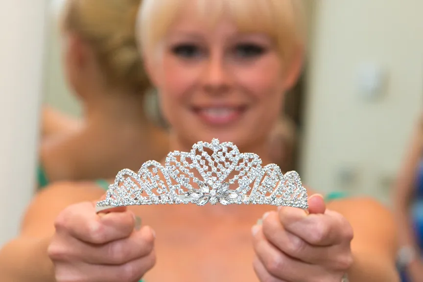 Woman smiling and holding a sparkling silver tiara toward the camera.