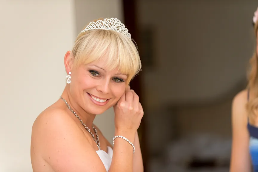 Blonde bride smiling and adjusting her earring, wearing a white dress and a tiara.