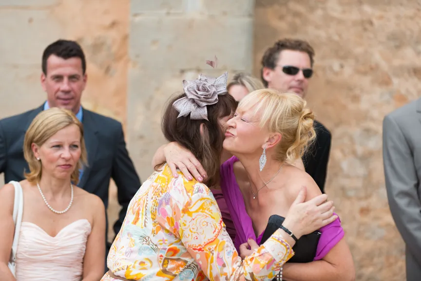 Two women warmly greeting each other with a hug and cheek kiss at an outdoor event with other attendees in the background.