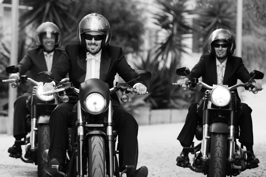 Three men in tuxedos riding motorcycles side by side on a paved street, all wearing helmets and sunglasses.