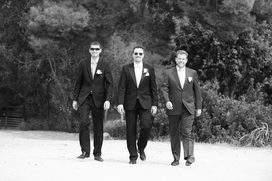 Three men in suits wearing boutonnieres walking outdoors with trees in the background, all smiling.