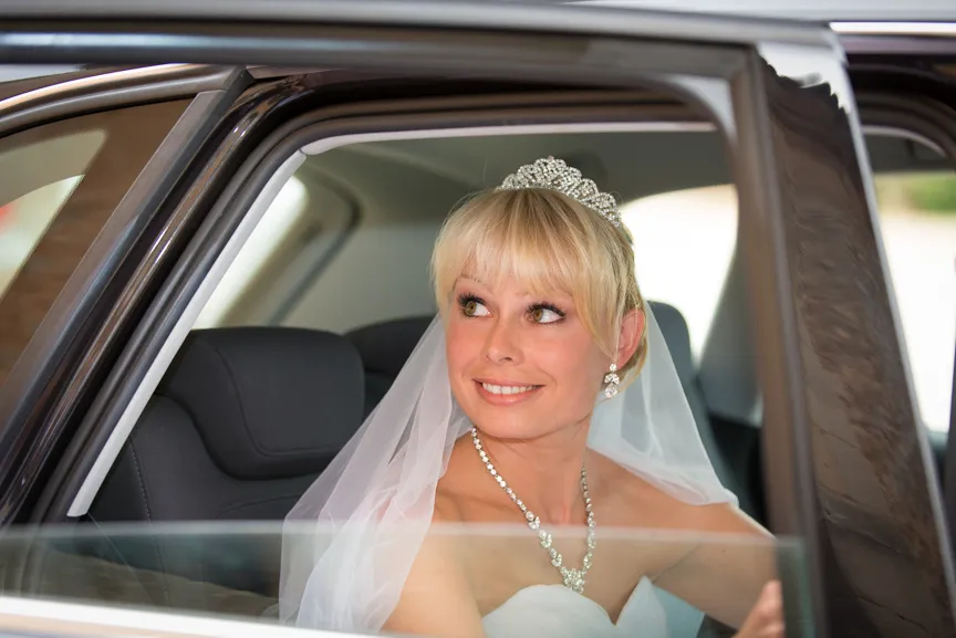 Smiling bride wearing a tiara, veil, and necklace sitting in the back seat of a car.