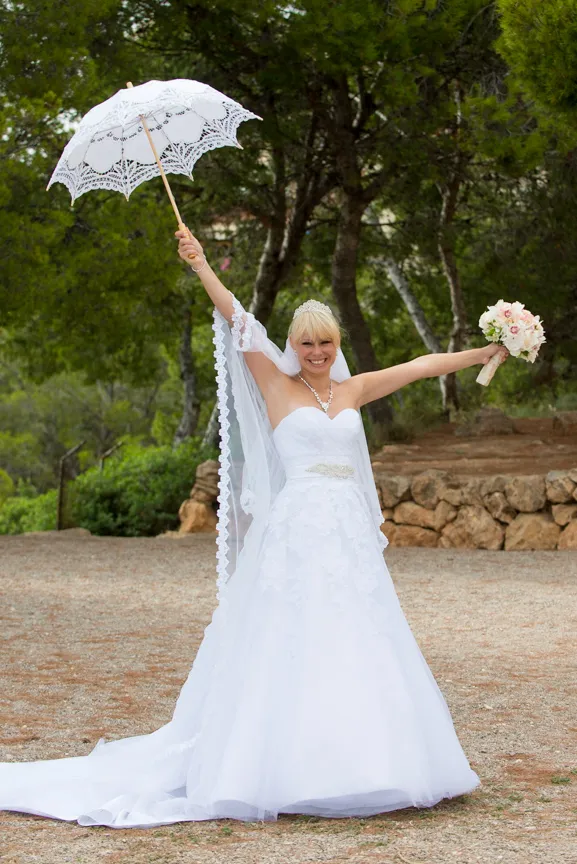 Smiling bride in a white wedding dress holding a lace parasol in one hand and a bouquet in the other, standing outdoors among trees.