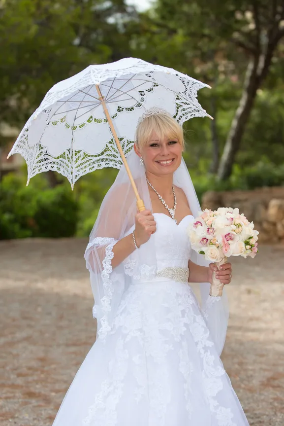 Smiling bride in a white lace wedding dress holding a bouquet of flowers and a white lace parasol outdoors.