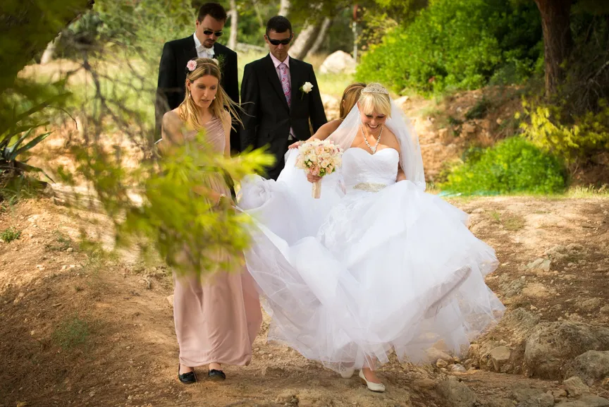 Bride in white wedding dress holding bouquet and lifting her gown walking outdoors with bridesmaid and two groomsmen on a rocky path.
