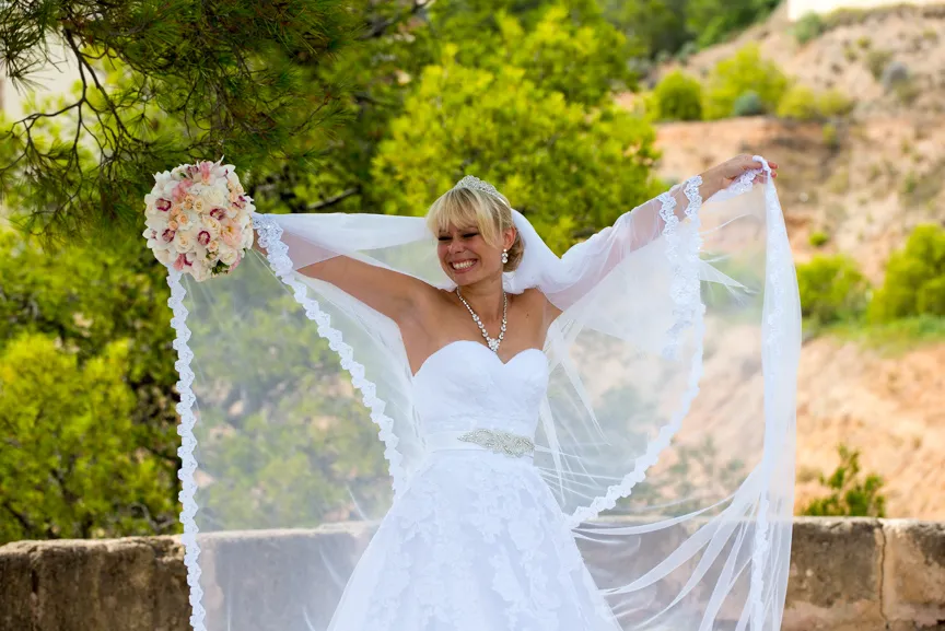 Smiling bride in a white wedding dress holds out her lace-trimmed veil outdoors with greenery and hills in the background.