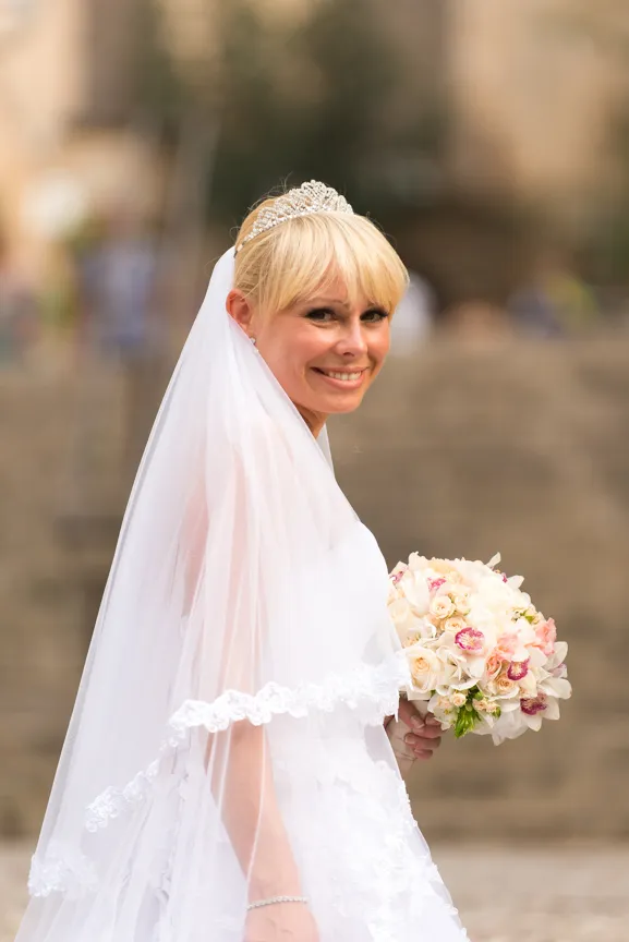 Smiling blonde bride wearing a tiara and lace veil, holding a bouquet of white and pink flowers.