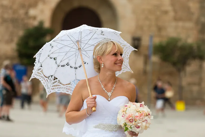 Bride in white dress holding lace parasol and bouquet, smiling and looking to the side in outdoor setting.