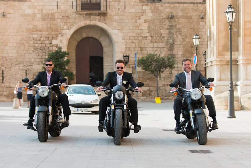 Three men in suits and sunglasses sitting on motorcycles in front of a historic stone building.