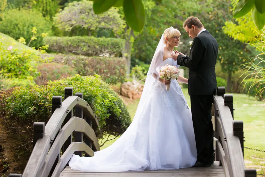 Bride in white wedding dress holding bouquet and groom in black suit standing on a wooden bridge surrounded by greenery.