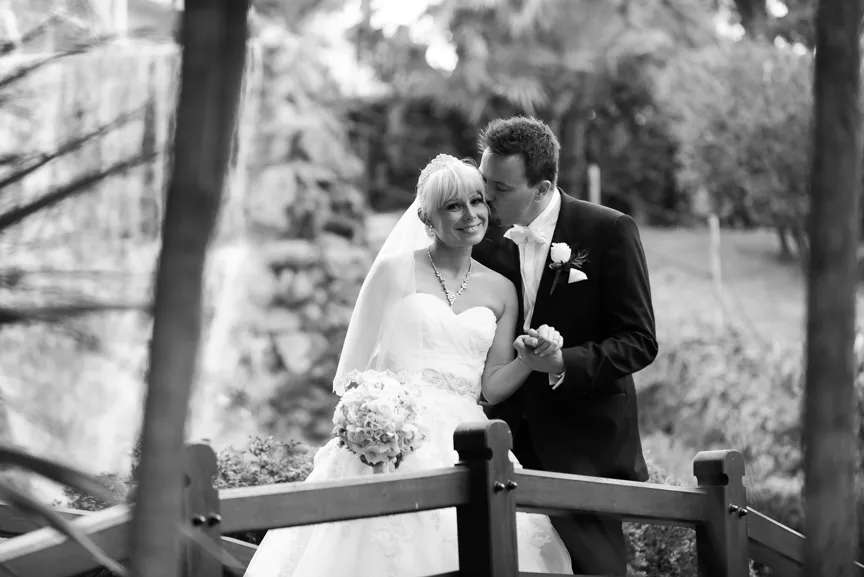 Bride in wedding gown holding a bouquet smiling as groom in tuxedo kisses her cheek on a wooden bridge in a garden.