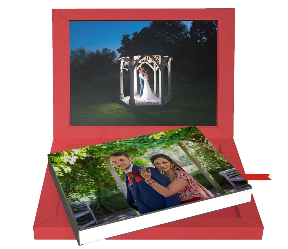 Red box containing a wedding photo book with an image of a smiling bride in traditional red attire hugging the groom in a blue suit, and a framed photo of the couple embracing under a gazebo at night.
