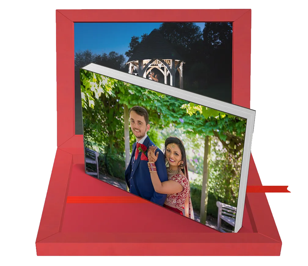 A red gift box contains a photo book open to a vibrant image of a smiling couple outdoors, with the woman in traditional attire holding the man from behind.