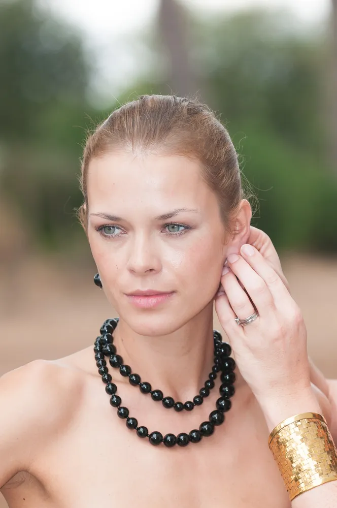 Woman with pulled-back hair wearing a double-strand black beaded necklace, adjusting an earring, and wearing a wide gold bracelet on her wrist outdoors.