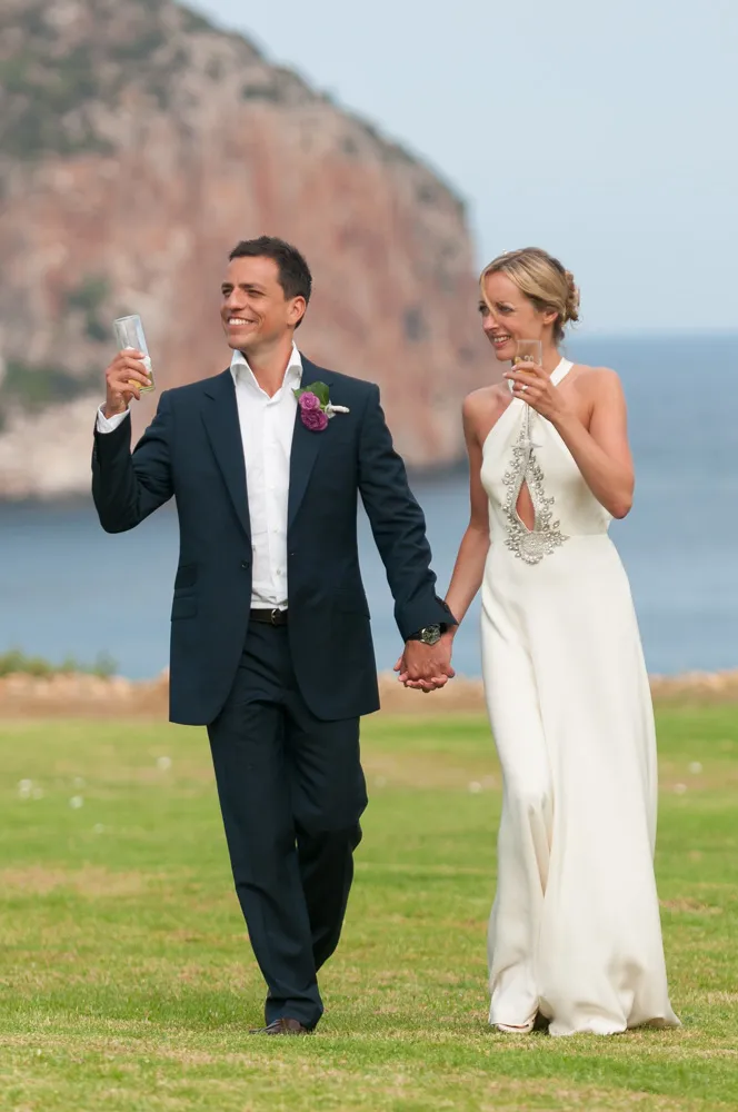 Bride and groom holding hands and toasting with glasses outdoors near a rocky coastline.