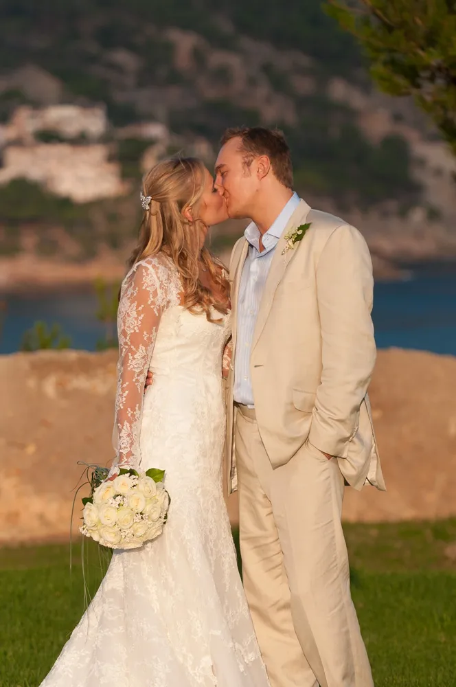 Bride in lace wedding dress holding bouquet kissing groom in light beige suit outdoors.