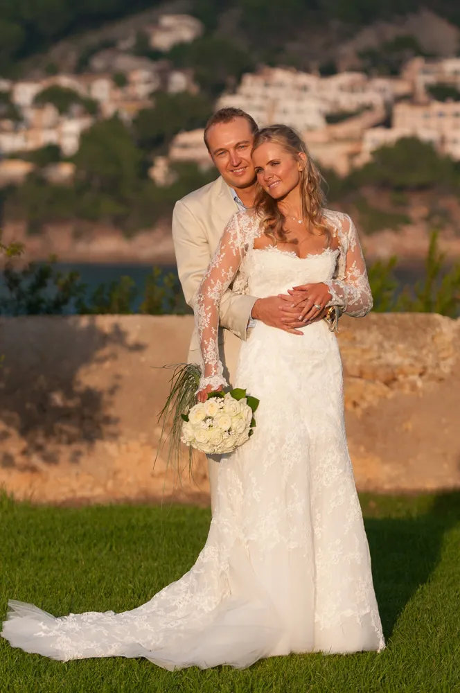 Bride in a long lace wedding dress holding a white rose bouquet, embraced by groom in a light-colored suit, standing outdoors on grass with blurred buildings and trees in the background.