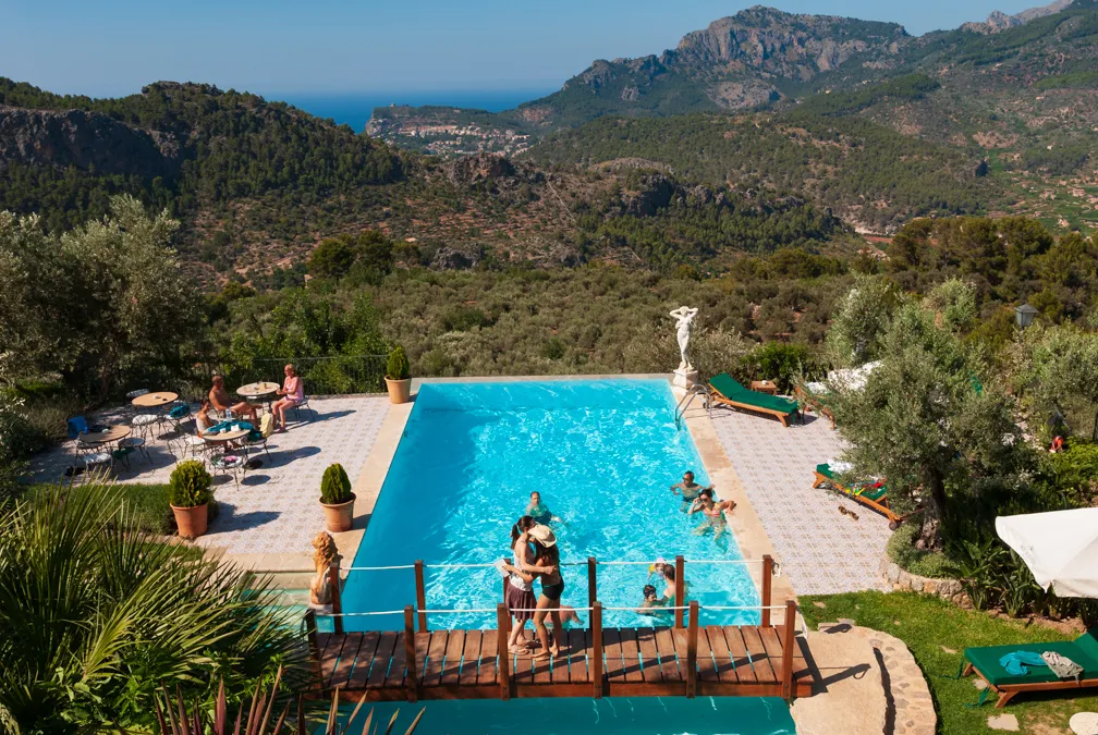 Outdoor pool surrounded by chairs and loungers with people swimming and embracing, set against a backdrop of forested mountains and clear sky.