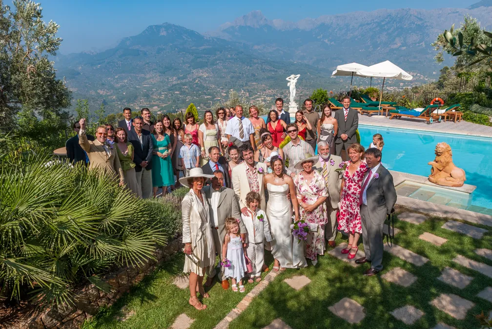 Group photo of wedding guests posing outdoors near a pool with mountain scenery in the background.
