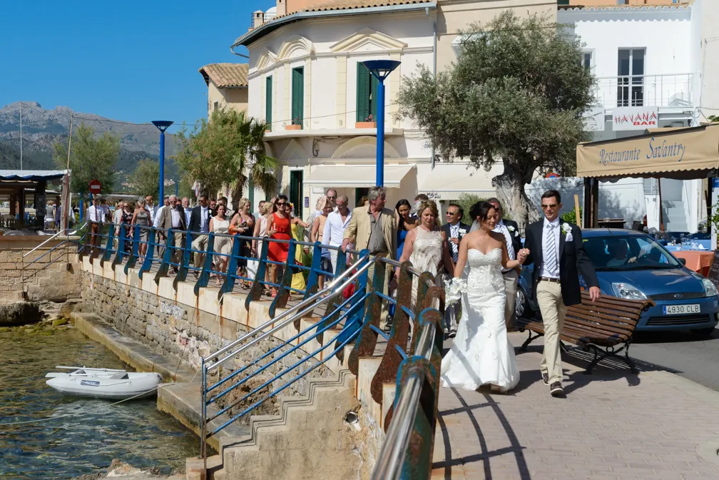 Bride and groom walking along a waterfront promenade followed by a large group of wedding guests on a sunny day.